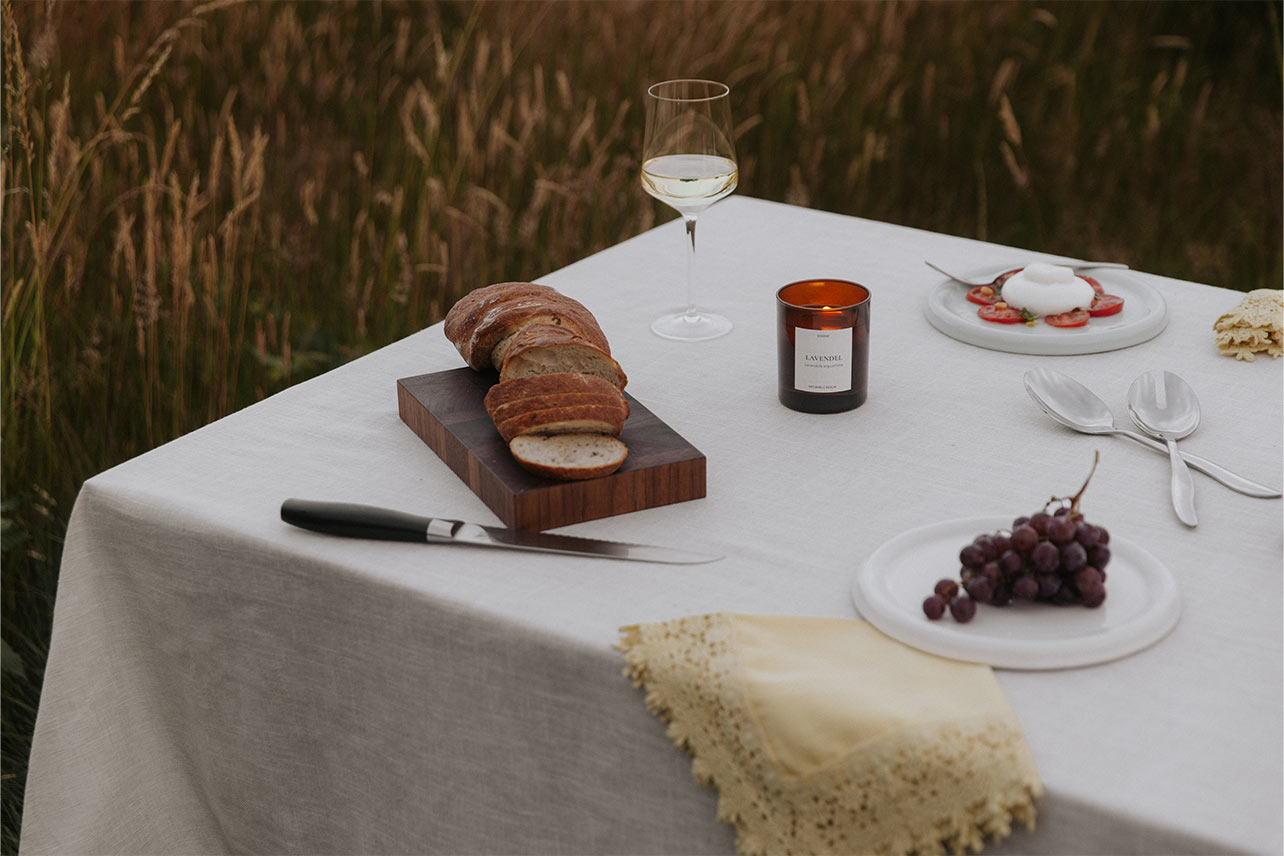Gedeckter Tisch im Sp&auml;tsommer mit Brot, Wein und der brennenden UpCandle Aromakerze Lavendel mit Holzdocht vor einem reifen Kornfeld.